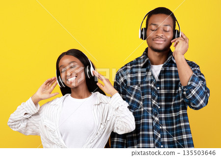 Favorite music during covid-19 lockdown at home. Happy teen african american students couple with closed eyes in modern wireless headphones have fun, isolated on yellow background, empty space 135503646
