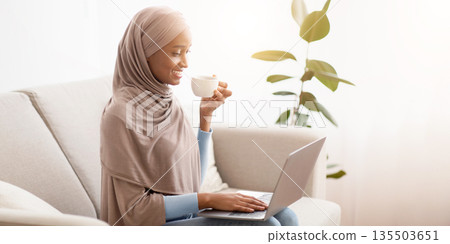Cheerful black lady in hijab sitting on couch with laptop and drinking coffee, indoors. Pretty African American woman with hot beverage having video call or broswing web on PC at home Cheerful black lady in hijab sitting on couch with laptop and drinking coffee, indoors. Pretty African American woman with hot beverage having video call or broswing web on PC at home 135503651