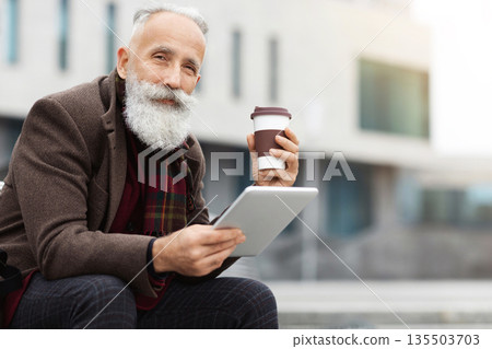 Handsome aged grey-haierd man holding paper cup with coffee and using modern digital tablet, sitting on street during break, copy space. Elderly man drinking morning coffee and using pad Handsome aged grey-haierd man holding paper cup with coffee and using modern digital tablet, sitting on street during break, copy space. Elderly man drinking morning coffee and using pad 135503703