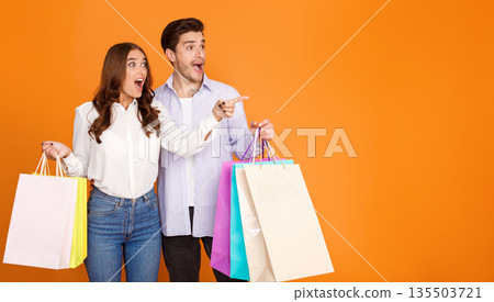 Wow sale. Shocked couple with shopping bags pointing fingers at free space, orange studio background 135503721