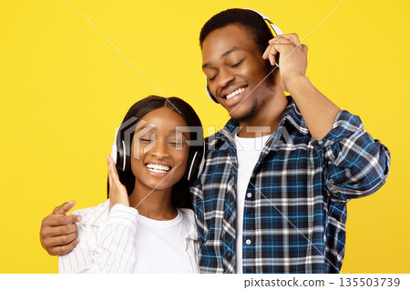 Favorite music together and stay home at covid-19. Smiling young african american couple in love in wireless headphones enjoy song at free time, isolated on yellow background, studio shot, copy space 135503739