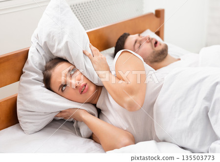 Husband Snoring Bothering Angry Wife Unable To Get To Sleep Covering Ears With Pillow Lying In Bed Indoor. Selective Focus Husband Snoring Bothering Angry Wife Unable To Get To Sleep Covering Ears With Pillow Lying In Bed Indoor. Selective Focus 135503773