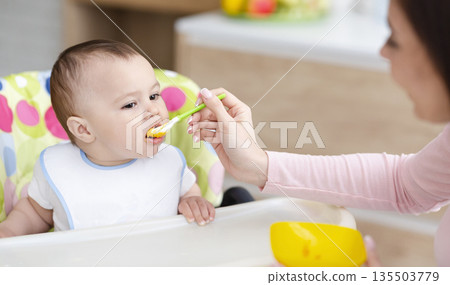 Mother feeding her cute toddler on high chair at kitchen, free space Mother feeding her cute toddler on high chair at kitchen, free space 135503779