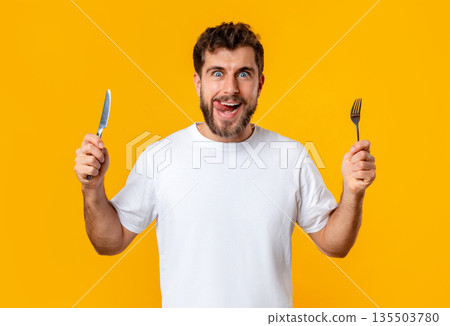 Hungry young guy holding knife and fork looking at camera, posing on yellow studio background. Portrait of funny young man having food cravings and temptation ready for tasty dinner 135503780