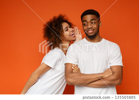Tell me your secrets. Pleasant smiling african-american woman whispering on ear to her man something important, orange background Tell me your secrets. Pleasant smiling african-american woman whispering on ear to her man something important, orange background 135503805