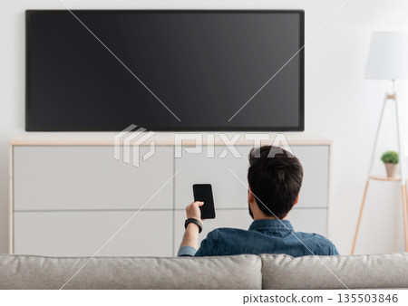Modern gadgets. Unrecognizable man with smartphone connected to TV, watching video at home, back view. Guy transfering video from phone to television. Information transfer between devices over wifi. Modern gadgets. Unrecognizable man with smartphone connected to TV, watching video at home, back view. Guy transfering video from phone to television. Information transfer between devices over wifi. 135503846