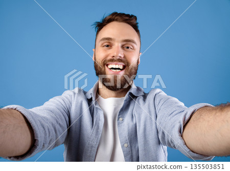Joyful bearded guy taking selfie on blue studio background. Positive millennial man making photo of himself, smiling at camera, having fun while doing mobile photography 135503851