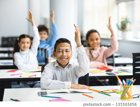 Education, Primary School, Learning and People Concept. Diverse group of smart junior schoolchildren sitting at desks in classroom and raising hands for an answer, studying with pleasure 135503856