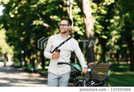 Healthy and active lifestyle, going to work on eco transport. Smiling attractive millennial male employee in glasses with bag and cup of takeaway coffee, moving bike in city park in summer, outdoor Healthy and active lifestyle, going to work on eco transport. Smiling attractive millennial male employee in glasses with bag and cup of takeaway coffee, moving bike in city park in summer, outdoor 135503866