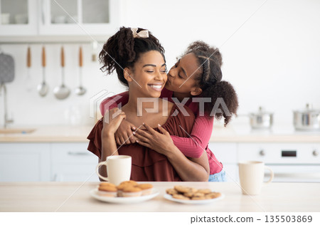 Cute black girl teenager hugging and kissing her happy beautiful mom after breakfast together, kitchen interior. Loving afro-american mother and daughter bonding while having snack, copy space 135503869