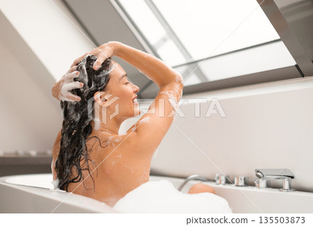 Haircare Cosmetics. Lady Applying Shampoo On Long Hair Washing Head Sitting In Bathtub Full Of Foam Indoors. Side View Shot Of Happy Woman Enjoying Hair Care Routine In Modern Bathroom Haircare Cosmetics. Lady Applying Shampoo On Long Hair Washing Head Sitting In Bathtub Full Of Foam Indoors. Side View Shot Of Happy Woman Enjoying Hair Care Routine In Modern Bathroom 135503873