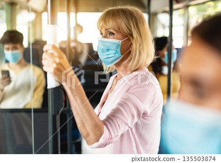 Safe Ride. Portrait of serious senior woman wearing protective face mask taking crowded bus holding handle, standing inside and looking out of window. Rush hour traffic, new normal life rules, covid Safe Ride. Portrait of serious senior woman wearing protective face mask taking crowded bus holding handle, standing inside and looking out of window. Rush hour traffic, new normal life rules, covid 135503874