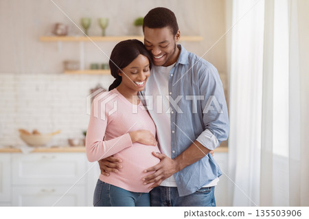 Black pregnant woman and her handsome husband embracing and smiling while spending time together near window at home. Romantic African American couple expecting new baby indoors. Parenthood concept Black pregnant woman and her handsome husband embracing and smiling while spending time together near window at home. Romantic African American couple expecting new baby indoors. Parenthood concept 135503906