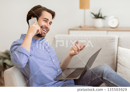 Cheerful Guy Talking On Phone Sitting With Laptop Computer On Sofa At Home. Modern Communication And Lifestyle. Happy Man Chatting On Cellphone And Laughing Indoors 135503929