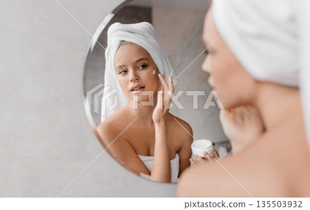 Home beauty routine. Young woman applying moisturising cream on face looking at miror in bathroom, free space. Attractive lady wearing white towel on head after shower 135503932
