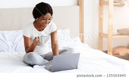 Happy young african american woman sitting on bed, websurfing on laptop at home, copy space. Smiling black lady looking at laptop screen, typing on keyboard, attending webinar, drinking coffee Happy young african american woman sitting on bed, websurfing on laptop at home, copy space. Smiling black lady looking at laptop screen, typing on keyboard, attending webinar, drinking coffee 135503945