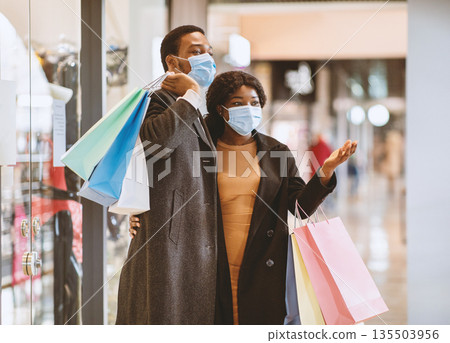 Seasonal sales during coronavirus pandemic. Happy african american family in coats and protective masks walking in mall after buying clothes at Black Friday, lady showing at store window, copy space 135503956