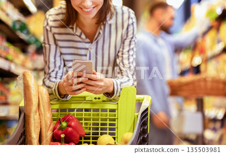 Couple In The Supermarket. Cropped closeup image of unrecognizable smiling woman leaning on shopping cart, using mobile phone, her boyfriend is choosing food in the blurred background Couple In The Supermarket. Cropped closeup image of unrecognizable smiling woman leaning on shopping cart, using mobile phone, her boyfriend is choosing food in the blurred background 135503981