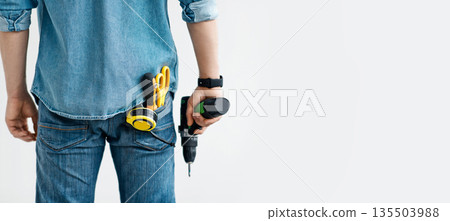 Men work, home renovation, construction or carpentry business. Young guy in jeans holds electric drill, with tools in his pocket, isolated on white background, panorama, empty space, cropped, back Men work, home renovation, construction or carpentry business. Young guy in jeans holds electric drill, with tools in his pocket, isolated on white background, panorama, empty space, cropped, back 135503988