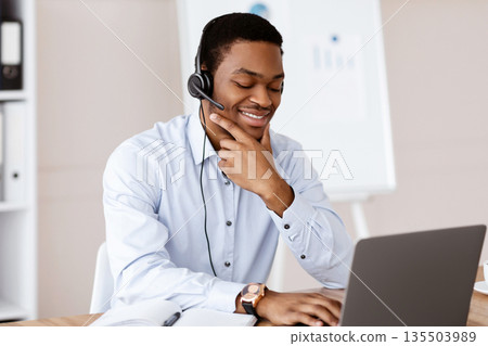 Positive young black man having online business conference in office, using laptop and headset, cheerfully talking to client or business partner, looking at computer screen and smiling, copy space Positive young black man having online business conference in office, using laptop and headset, cheerfully talking to client or business partner, looking at computer screen and smiling, copy space 135503989