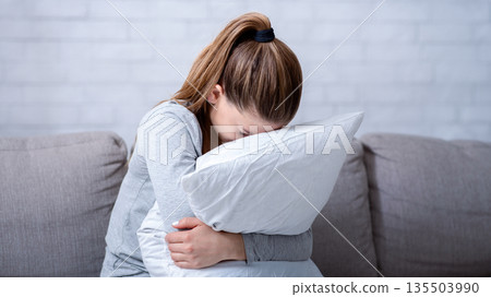 Young woman with depression hugging pillow and crying on sofa at home. Lonely millennial lady feeling stressed and hopeless, suffering from mood disorder or having psychological problem Young woman with depression hugging pillow and crying on sofa at home. Lonely millennial lady feeling stressed and hopeless, suffering from mood disorder or having psychological problem 135503990