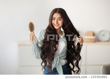 Portrait of happy Indian lady brushing her long dark hair with wooden brush at home. Lovely young woman taking care of her beautiful locks. Beauty, wellness and hairdressing concept Portrait of happy Indian lady brushing her long dark hair with wooden brush at home. Lovely young woman taking care of her beautiful locks. Beauty, wellness and hairdressing concept 135503991