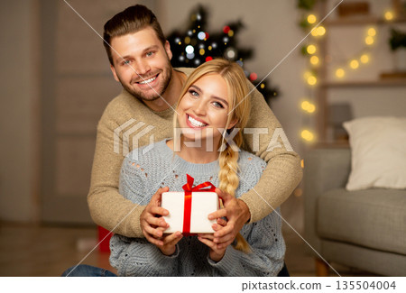 Cheerful young couple hugging and holding gift box, exchanging with Christmas presents at home, empty space. Smiling man and woman presenting xmas gifts to each other, celebrating winter holidays 135504004
