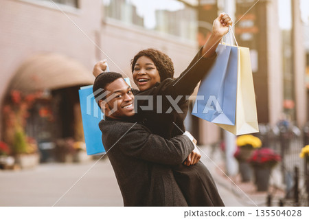 Good bought at low price during seasonal sale and Black Friday. Happy millennial man in coat picks up wife with colored bags and rejoice successful purchase, near modern city mall, free space Good bought at low price during seasonal sale and Black Friday. Happy millennial man in coat picks up wife with colored bags and rejoice successful purchase, near modern city mall, free space 135504028