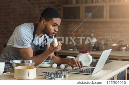 Black man baking pastry and looking recipe on laptop in loft kitchen. 135504030