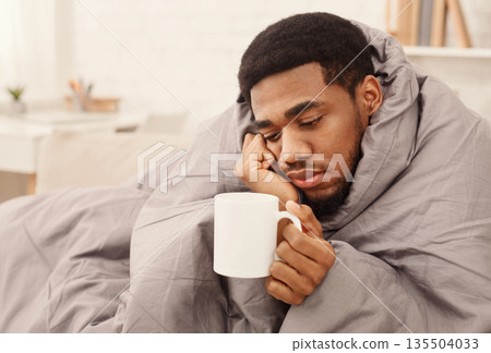 Sad african-american man with flu wrapped in warm blanket, holding cup with tea, copy space Sad african-american man with flu wrapped in warm blanket, holding cup with tea, copy space 135504033