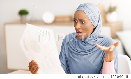 Angry black woman in hijab finding mistake in document near laptop computer at home. Young lady in traditional headscarf displeased about too much paperwork, indoors. Remote job concept 135504046