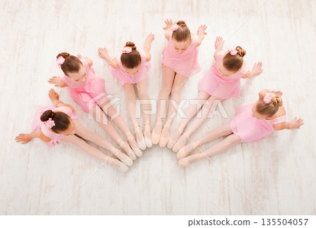 Little ballerinas in ballet studio, top view. Group of girls practicing position sitting on floor, classical dance studio Little ballerinas in ballet studio, top view. Group of girls practicing position sitting on floor, classical dance studio 135504057