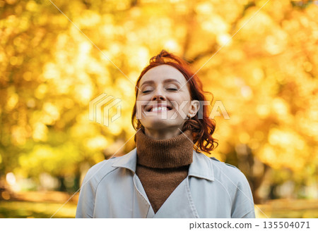 Satisfied caucasian millennial red-haired female in raincoat enjoys warm autumn and freedom, breathes fresh air in park with yellow leaves. Rest and relax alone in city at end of covid-19 quarantine 135504071