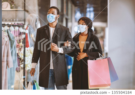 Sale, consumerism, Black friday and shopping during coronavirus epidemic. Young african american loving couple in protective mask with shopping bags walking in mall and looking at showcases in store 135504184