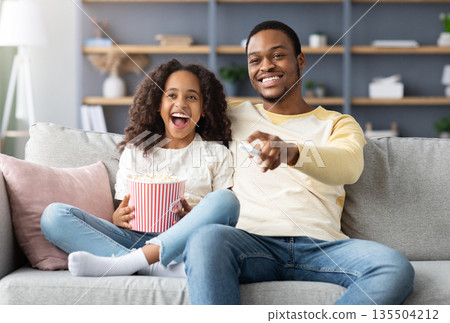 Happy black family father and daughter watching movie together at home. Laughing african american dad and school girl watching funny cartoon and eating popcorn, sitting on couch, copy space 135504212