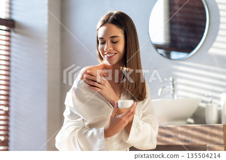 Body Care Concept. Young Beautiful Woman Applying Moisturising Cream On Shoulder In Bathroom, Smiling Attractive Lady Wearing White Silk Robe Making Beauty Treatments At Home, Free Space Body Care Concept. Young Beautiful Woman Applying Moisturising Cream On Shoulder In Bathroom, Smiling Attractive Lady Wearing White Silk Robe Making Beauty Treatments At Home, Free Space 135504214