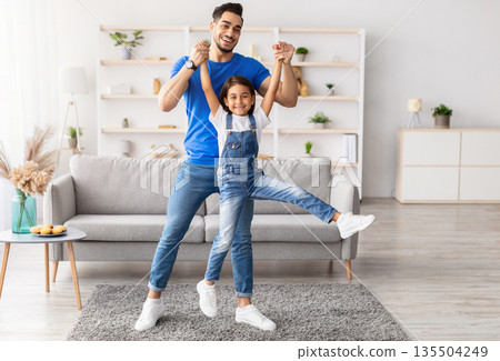 Happy Loving Family. Full body length of young smiling dad spending free time together with his cute little daughter in living room at home, holding her hands and lifting up, having fun and jumping Happy Loving Family. Full body length of young smiling dad spending free time together with his cute little daughter in living room at home, holding her hands and lifting up, having fun and jumping 135504249