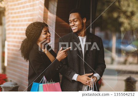 Enjoy shopping on black friday or season sale. Cheerful millennial african american lady with many colored paper bags in hands shows to guy at window and calls to store, free space Enjoy shopping on black friday or season sale. Cheerful millennial african american lady with many colored paper bags in hands shows to guy at window and calls to store, free space 135504252