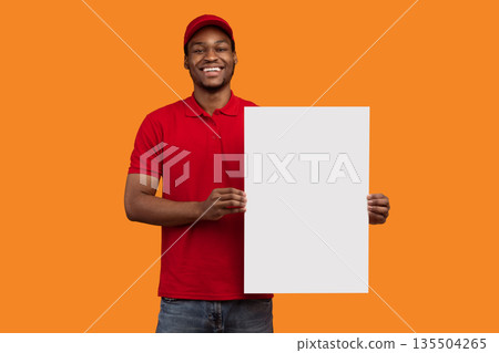 Shipment Service Promotion. Portrait of cheerful bearded black delivery man in red uniform holding and showing vertical empty blank board sign standing isolated on orange studio background 135504265