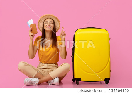 Happy young woman in causal outfit with passport and flight tickets sitting by yellow suitcase, closing eyes and crossing fingers, waiting for vacation, pink studio background, copy space Happy young woman in causal outfit with passport and flight tickets sitting by yellow suitcase, closing eyes and crossing fingers, waiting for vacation, pink studio background, copy space 135504300