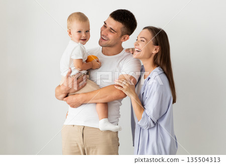 Happy Family Of Three With Cute Infant Baby Posing Over White Background, Cheerful Young Parents And Their Adorable Little Child Embracing And Laughing, Enjoying Spending Time Together, Copy Space Happy Family Of Three With Cute Infant Baby Posing Over White Background, Cheerful Young Parents And Their Adorable Little Child Embracing And Laughing, Enjoying Spending Time Together, Copy Space 135504313