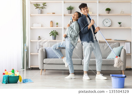 Happy asian couple singing songs and dancing while cleaning living room, using broom and mop as microphones, cheerful young man and woman imitating rock stars while house-keeping, copy space Happy asian couple singing songs and dancing while cleaning living room, using broom and mop as microphones, cheerful young man and woman imitating rock stars while house-keeping, copy space 135504329