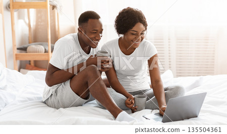 Joyful african american family in pajamas sitting on bed, watching movie on laptop, home interior, copy space. Happy black man and woman spending weekend together, drinking tea and websurfing 135504361