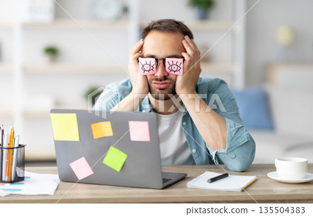 Tired young man having stickers with open eyes drawing on his glasses, sleeping in front of laptop at home office. Exhausted millennial man overworking, napping, failing to meet deadline 135504383