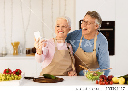 Joyful Elderly Spouses Making Selfie On Mobile Phone Having Fun Cooking Together Standing In Kitchen At Home. Modern Retirement Lifestyle, Senior People Using Gadgets And Enjoying Life Concept Joyful Elderly Spouses Making Selfie On Mobile Phone Having Fun Cooking Together Standing In Kitchen At Home. Modern Retirement Lifestyle, Senior People Using Gadgets And Enjoying Life Concept 135504417