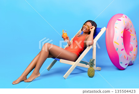 Full length portrait of young black woman chilling in lounge chair with yummy tropical cocktail on blue studio background. Relaxed millennial lady in swimwear enjoying her summer vacation 135504423