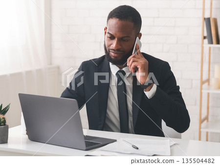Serious african american entrepreneur having phone conversation with business partners, using laptop, office interior, copy space Serious african american entrepreneur having phone conversation with business partners, using laptop, office interior, copy space 135504482
