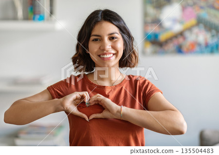 Smiling Young Arab Woman Showing Heart Gesture With Hands Near Chest, Happy Middle Eastern Millennial Lady Making Love Sign At Camera While Standing Indoors In Home Interior, Copy Space 135504483