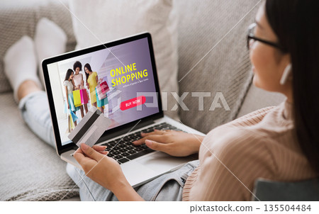Online Shopping Concept. Back view of asian girl using laptop, looking at website, holding credit card, sitting at home Online Shopping Concept. Back view of asian girl using laptop, looking at website, holding credit card, sitting at home 135504484