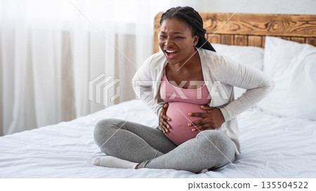 Pregnant black lady having labor contractions at home, suffering from acute belly ache and painful abdominal cramps, sitting on bed and touching her pregnant tummy, feeling unwell, free space Pregnant black lady having labor contractions at home, suffering from acute belly ache and painful abdominal cramps, sitting on bed and touching her pregnant tummy, feeling unwell, free space 135504522